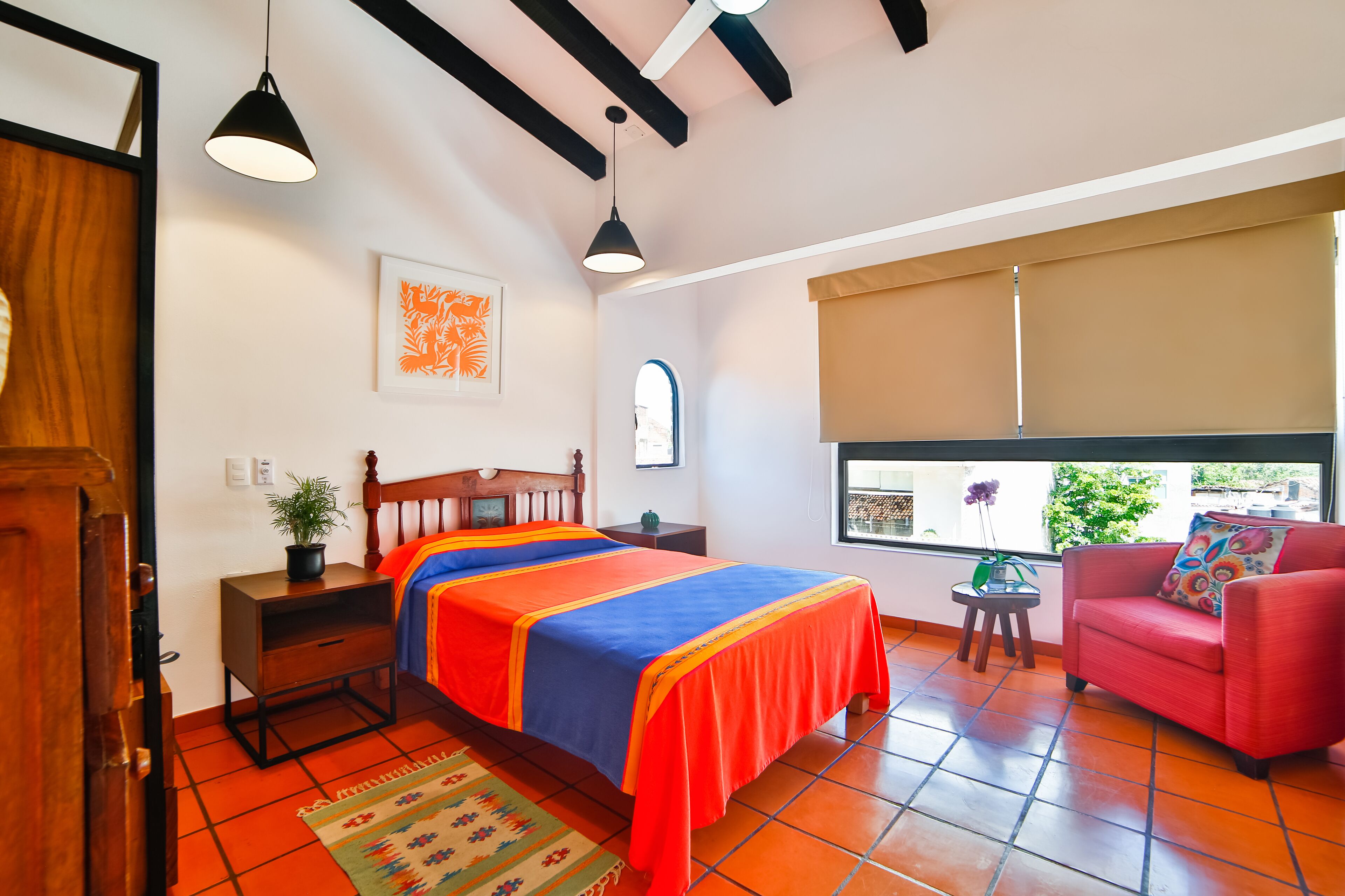 A bedroom with a bed, bedside table, potted plant, window with blinds, and a red chair.