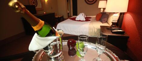 Une chambre d’hôtel comprenant un lit, une table de chevet avec une lampe, un minibar et un plateau avec une bouteille de champagne, des verres et des fruits.
