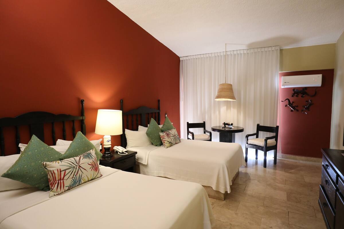 A hotel room with two beds, a desk, chairs, a lamp, and an air conditioning unit.