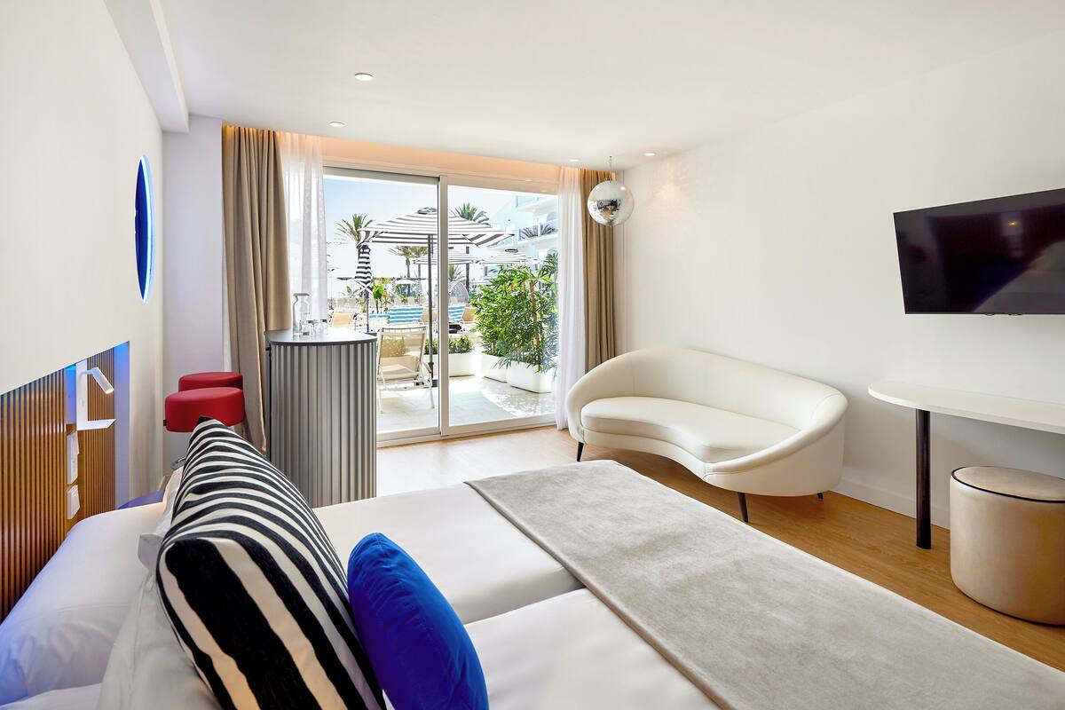 A modern hotel room with a bed, a sofa, a TV, and a view of the pool area.