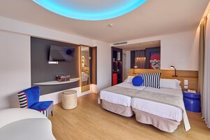 Una habitación de hotel moderna con una cama grande, un televisor montado en la pared, un área de descanso con una silla y una mesita.