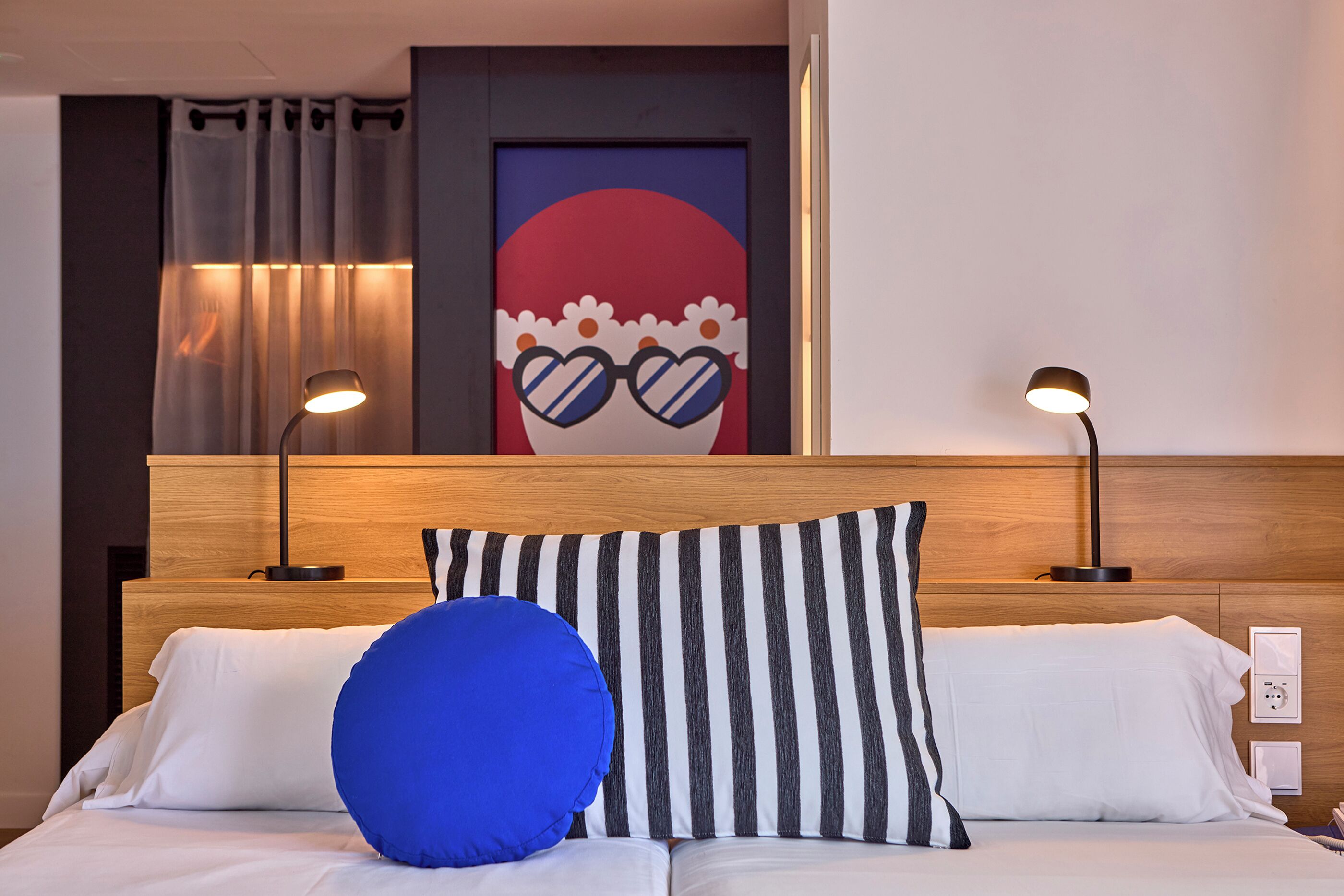 Habitación de hotel con cama, dos lámparas de noche, una obra de arte en la pared y una cortina.