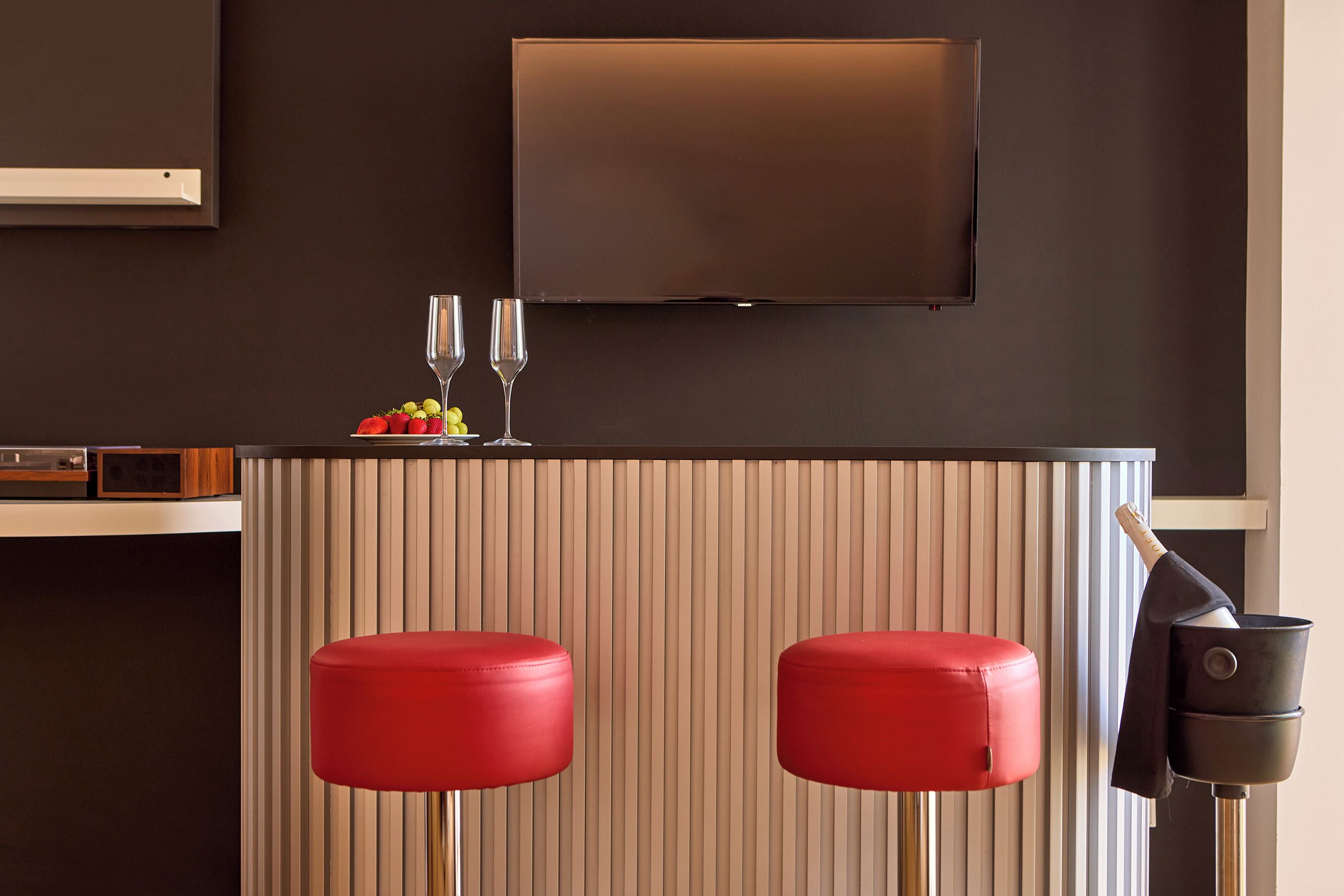 A modern bar area with two red stools, a flat-screen TV mounted on the wall, and a wine bottle on a stand.