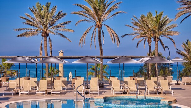 Zona junto a la piscina con sillones de descanso y sombrillas, palmeras y vista al océano.