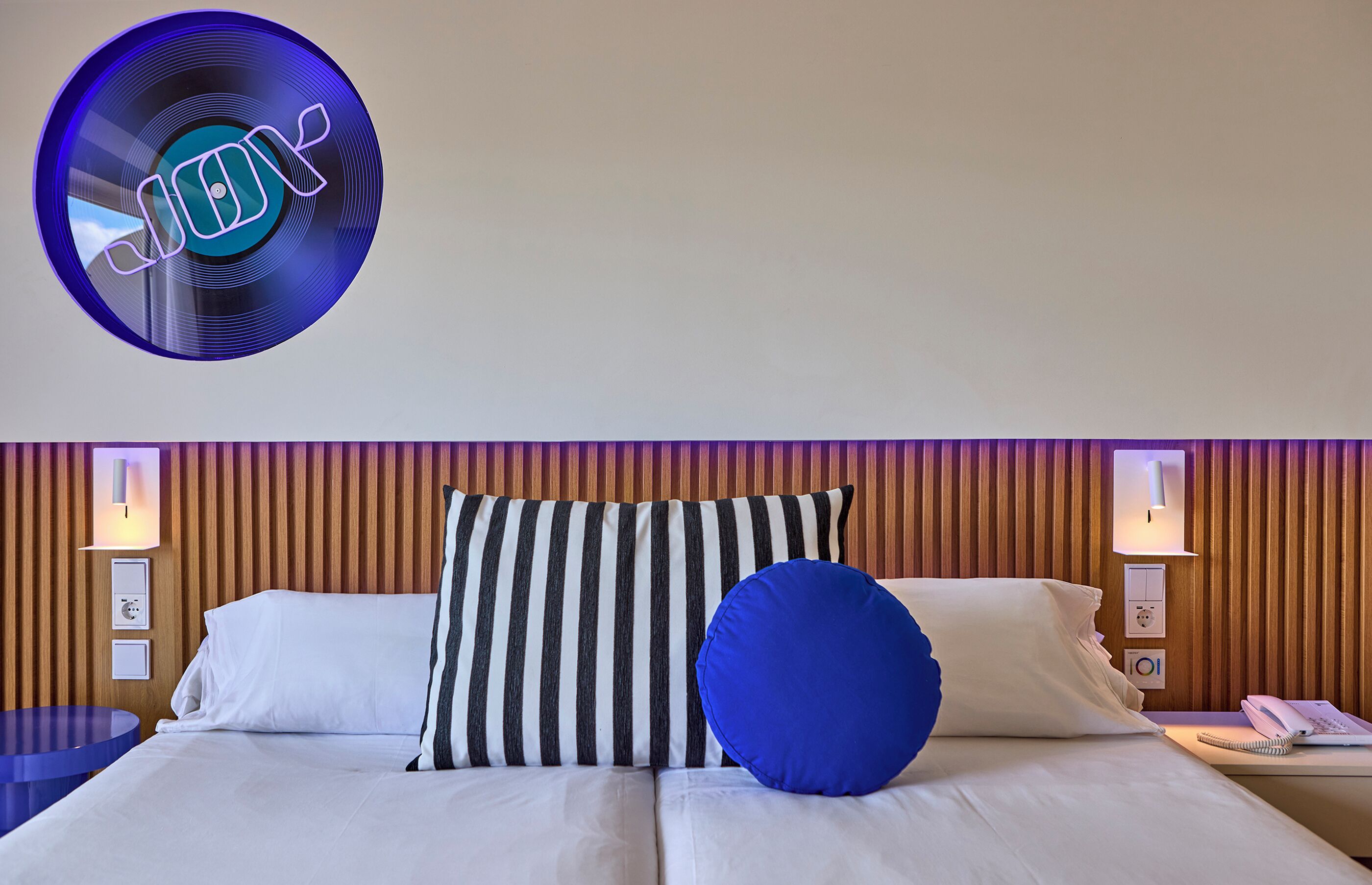 A hotel room with a bed, striped pillows, a blue pillow, and a wall-mounted light fixture.