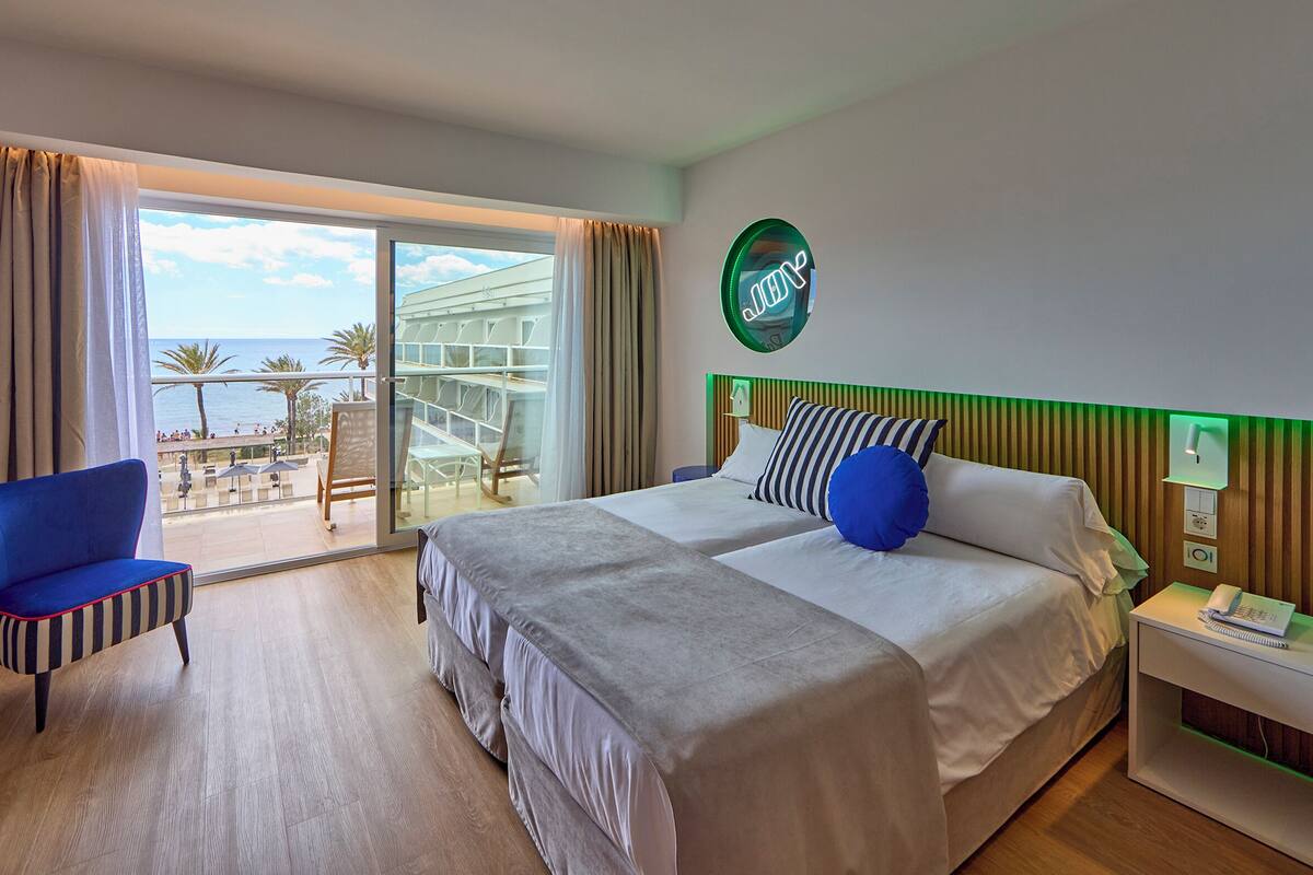 Habitación de hotel con una cama grande, una silla, una mesita de noche y vista al océano a través de una puerta corrediza de vidrio.