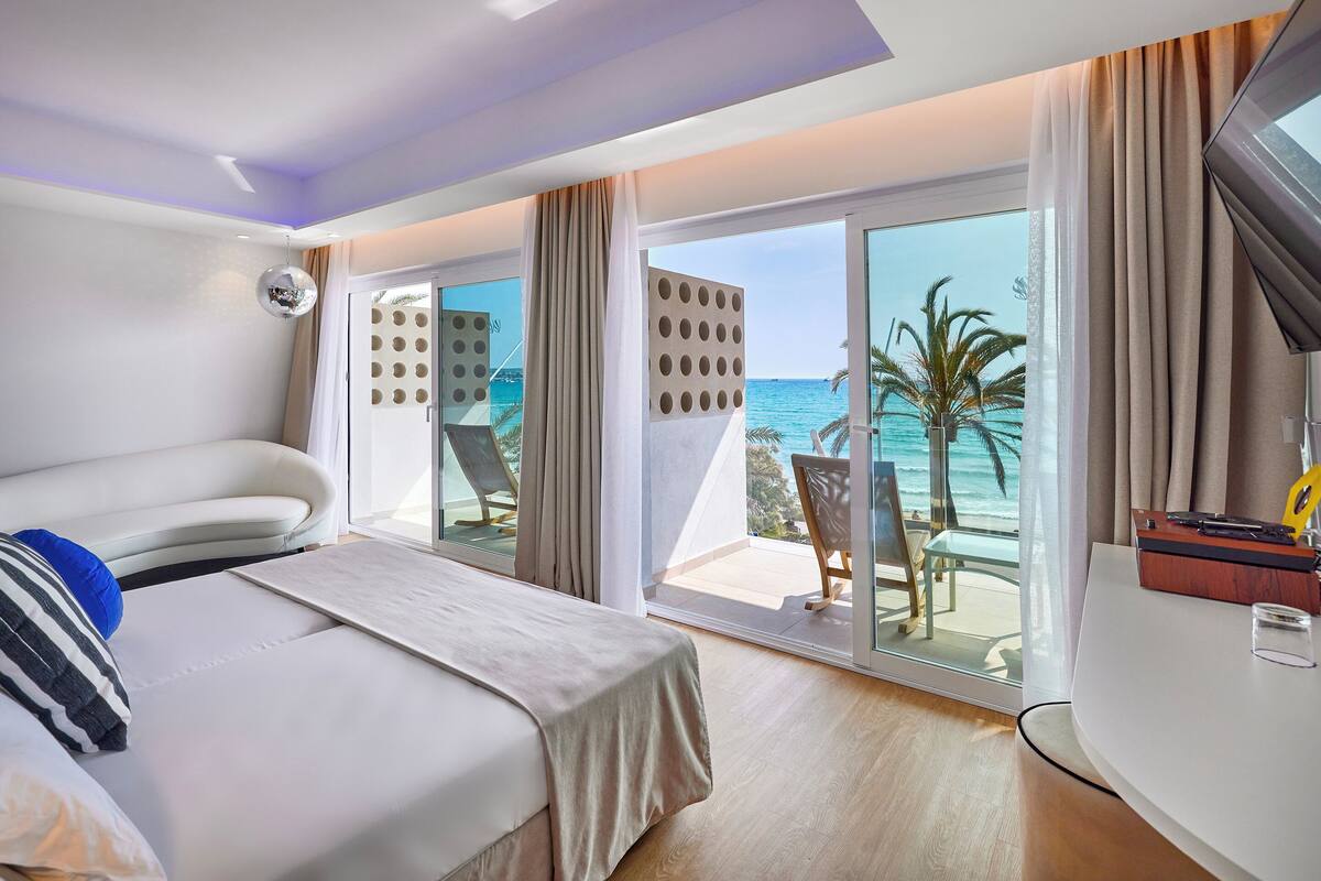 A hotel room with a bed, a TV, a desk, and a balcony with a view of the sea.