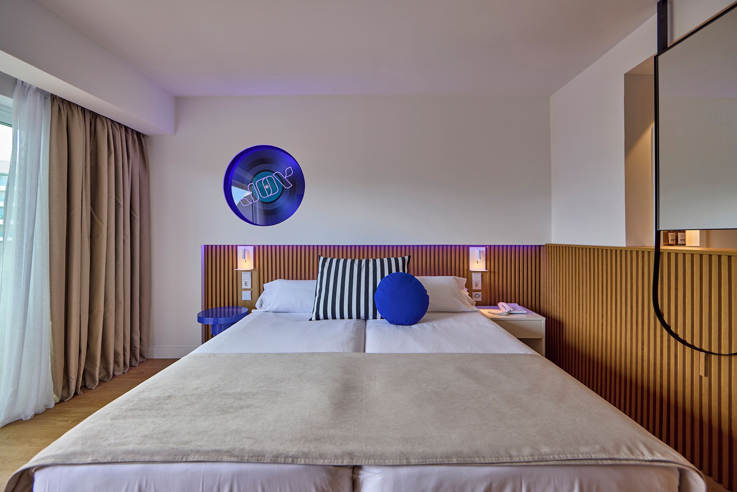 A hotel room with a large bed, a round wall clock, a bedside table with a lamp, and a wooden panel wall.