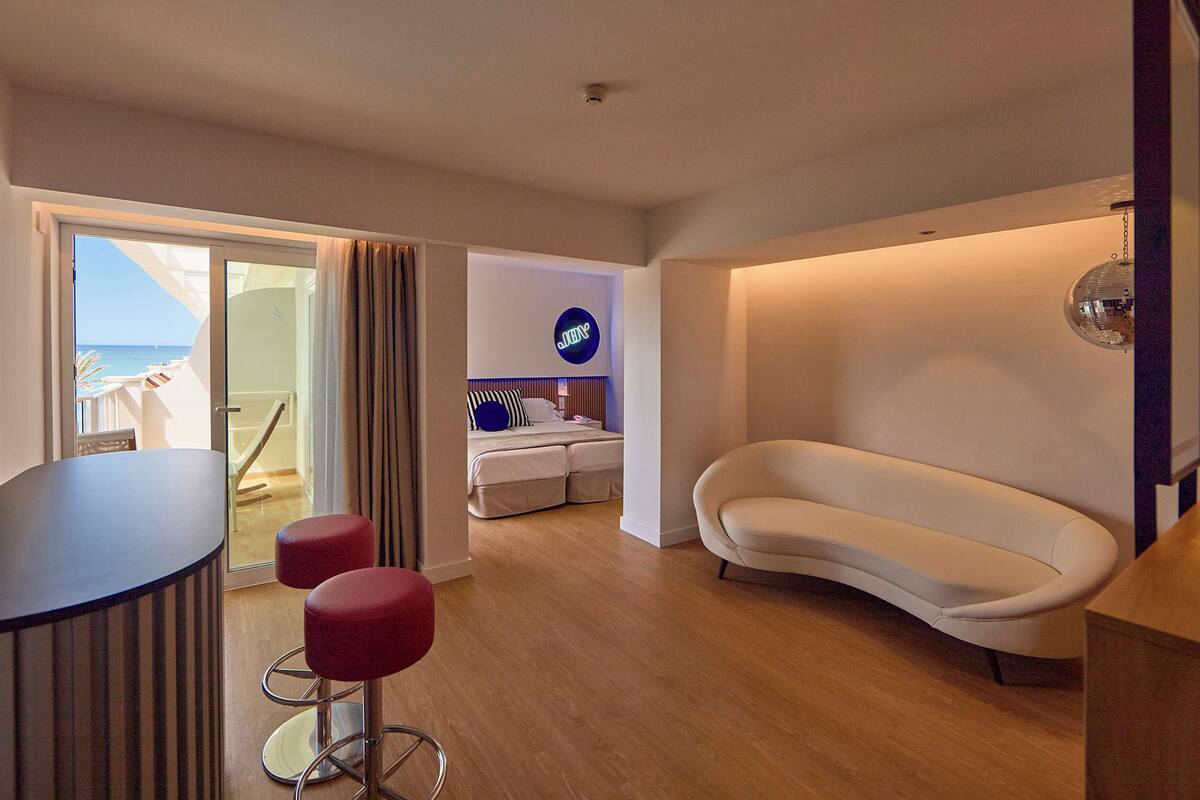 A modern hotel room with a balcony, a sofa, a bar area, and a bed with a neon sign.