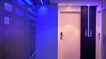 Un pasillo con una pared iluminada en azul, una puerta blanca y un ascensor negro con los números 107 y 106.
