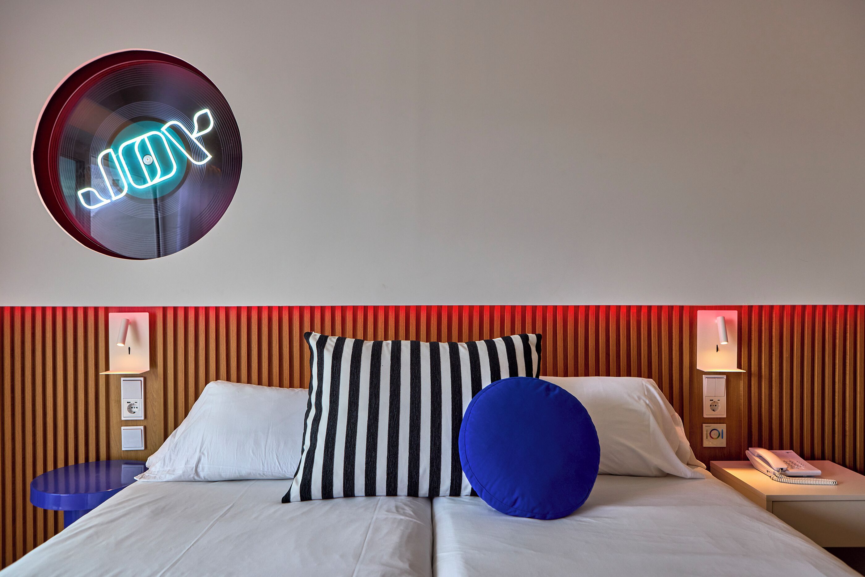 A hotel room with a bed, striped pillows, a blue pillow, a neon sign, a bedside table, and a nightstand with a phone and a lamp.