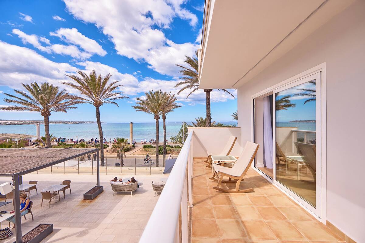 A balcony with a view of the beach, palm trees, and a pool area with sun loungers and tables.