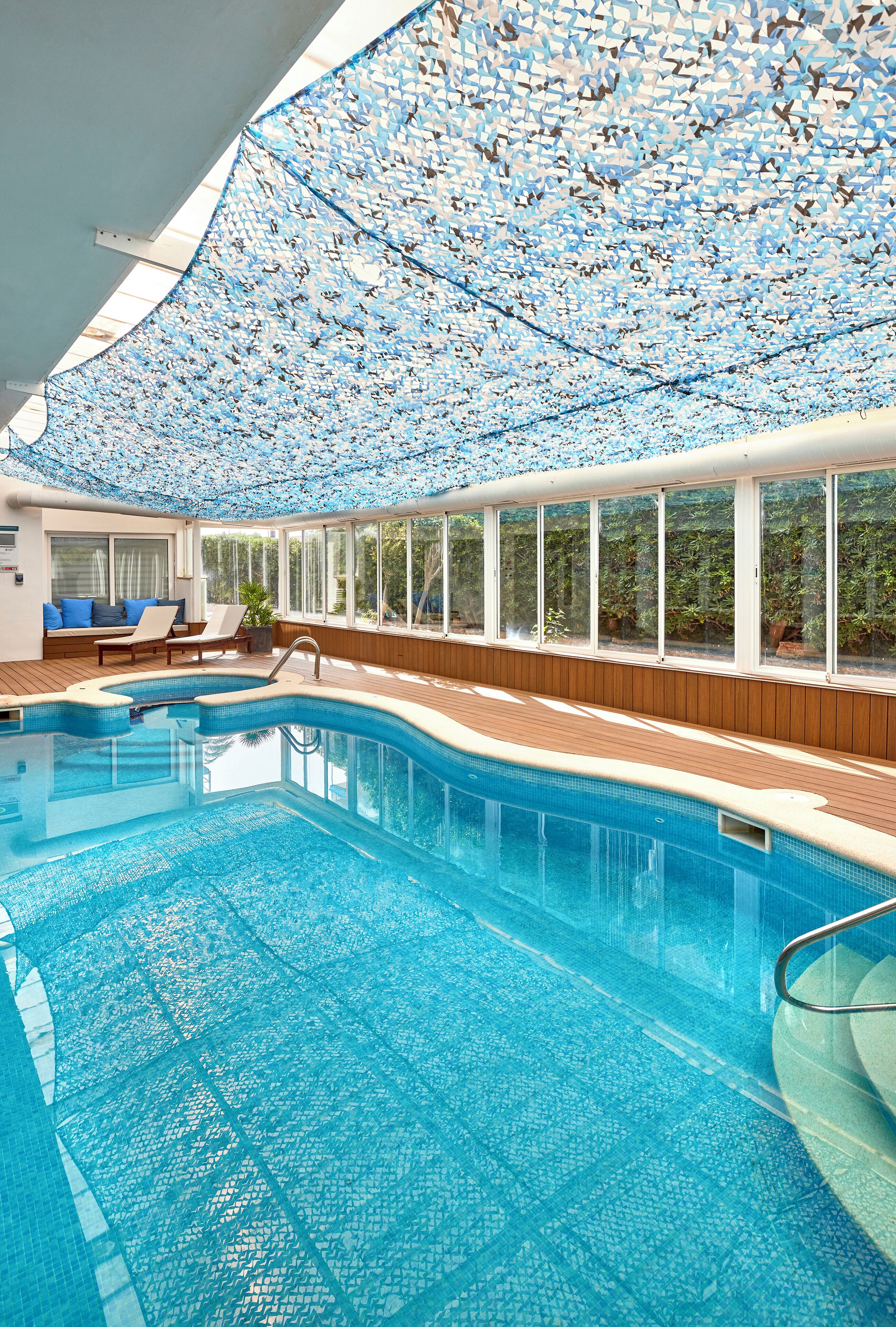 Une piscine intérieure dotée d’une terrasse en bois, de chaises longues et d’un plafond orné de motifs décoratifs.