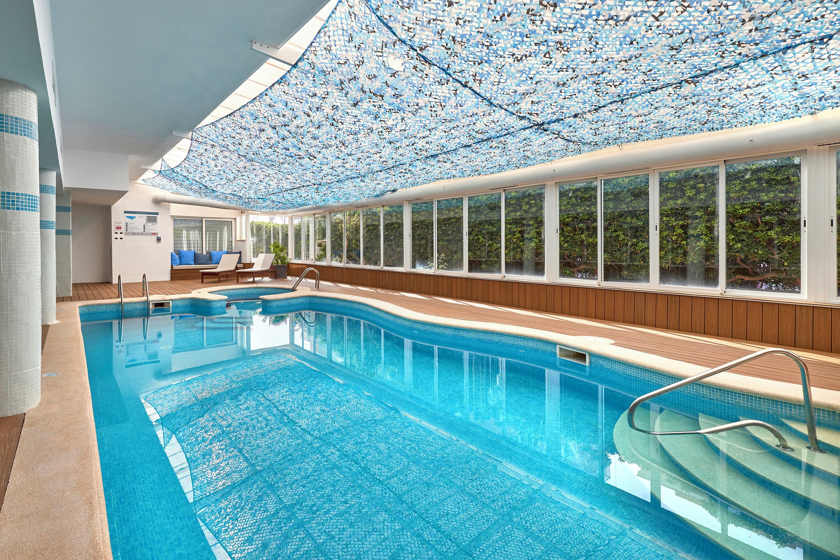An indoor swimming pool with clear blue water, a wooden deck, and a ceiling covered with a patterned fabric.