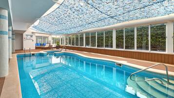 An indoor swimming pool with clear blue water, a wooden deck, and a ceiling covered with a patterned fabric.