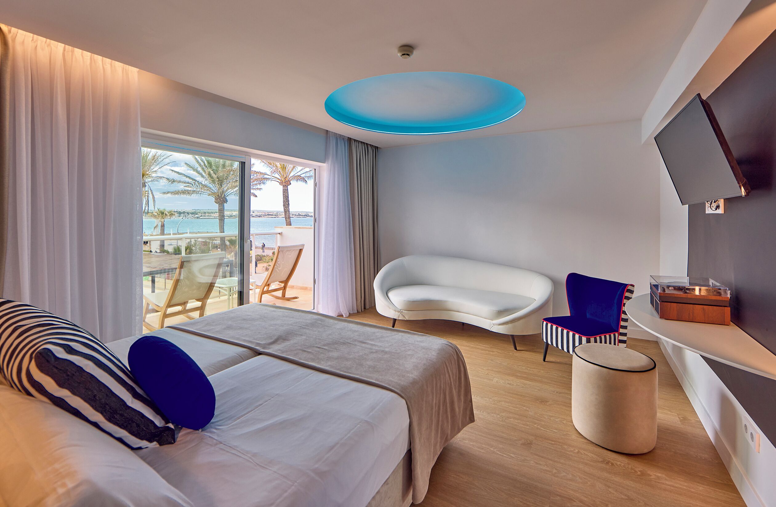 Una habitación de hotel moderna con una cama grande, una butaca blanca curvada, un pequeño puf redondo y un televisor montado en la pared.