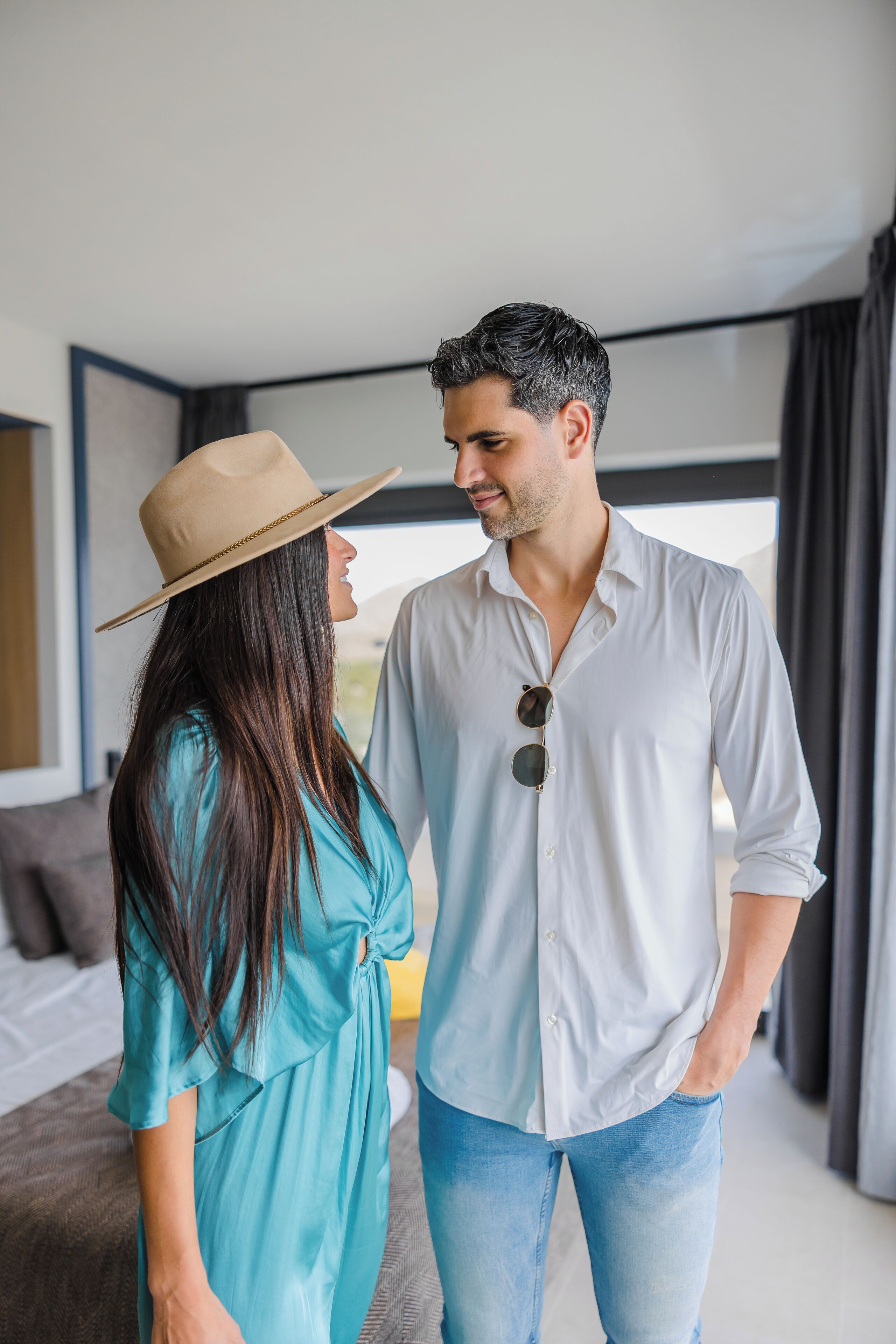 Una pareja en una habitación de hotel moderna con una cama grande, un ventanal con cortinas y vistas al exterior.