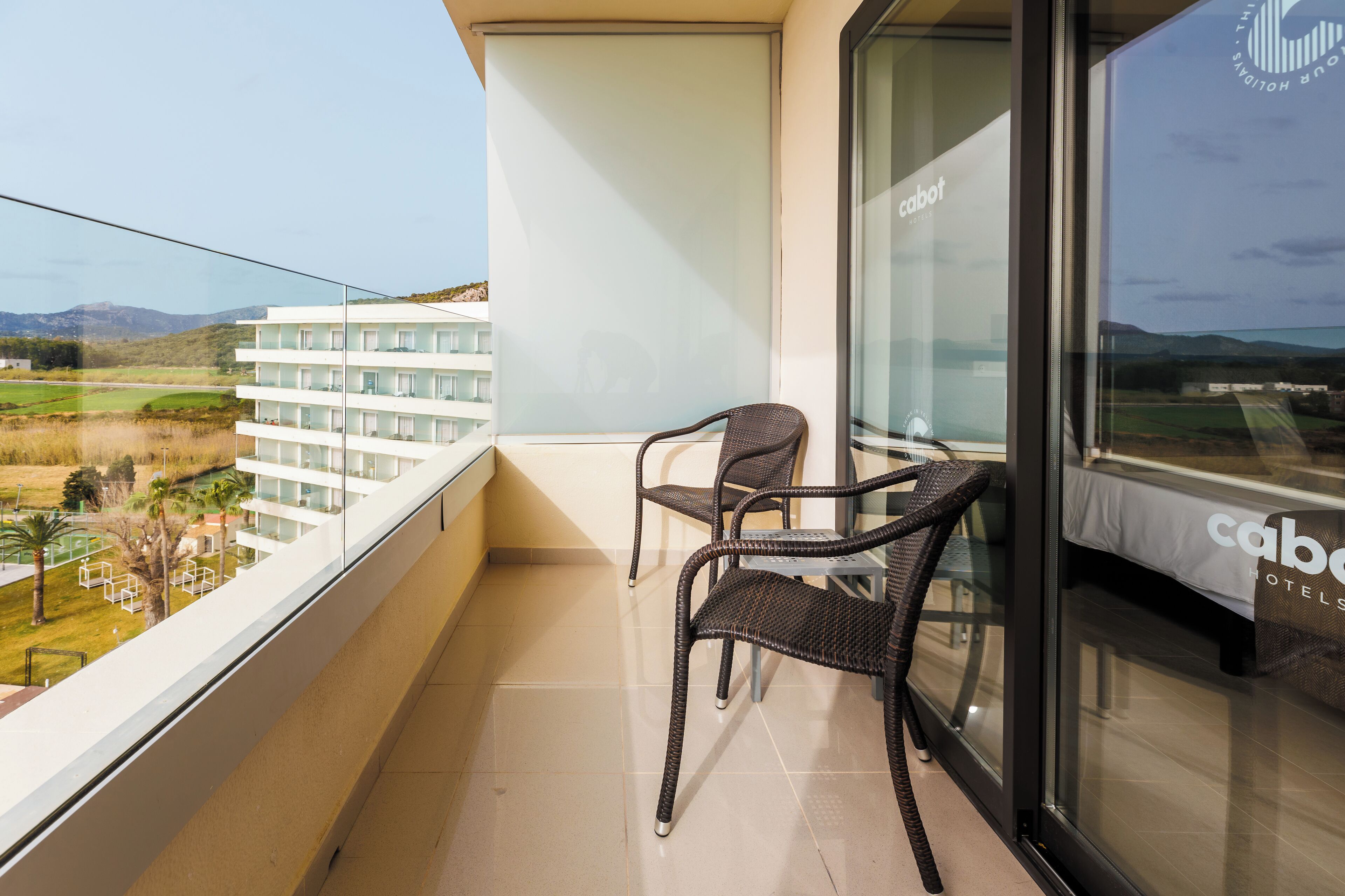 A balcony with wicker chairs, a glass table, and a view of a golf course and buildings.