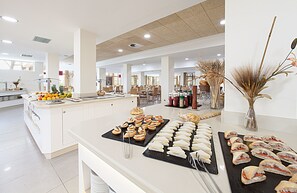 Un bufé con diversas opciones de comida, como sándwiches y pasteles, en un amplio comedor con decoración blanca y múltiples disposiciones de mesas.