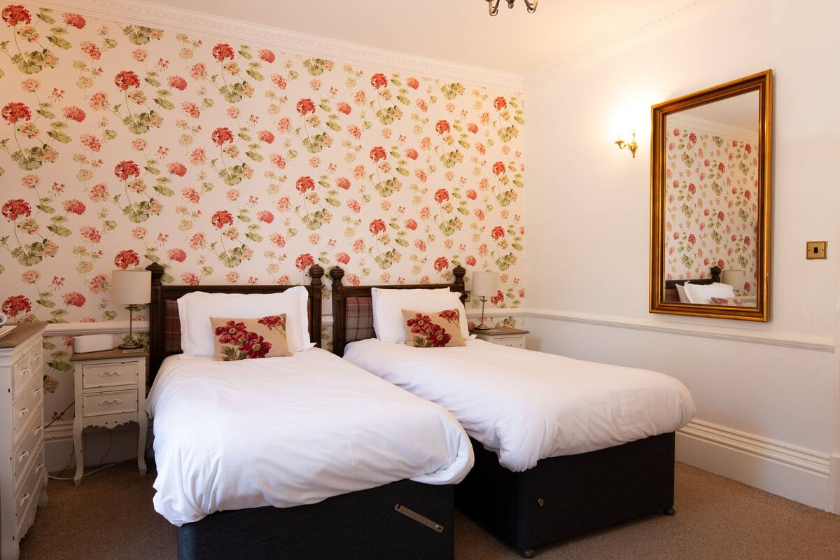 Un dormitorio con dos camas, papel tapiz floral, un espejo y un aparador.
