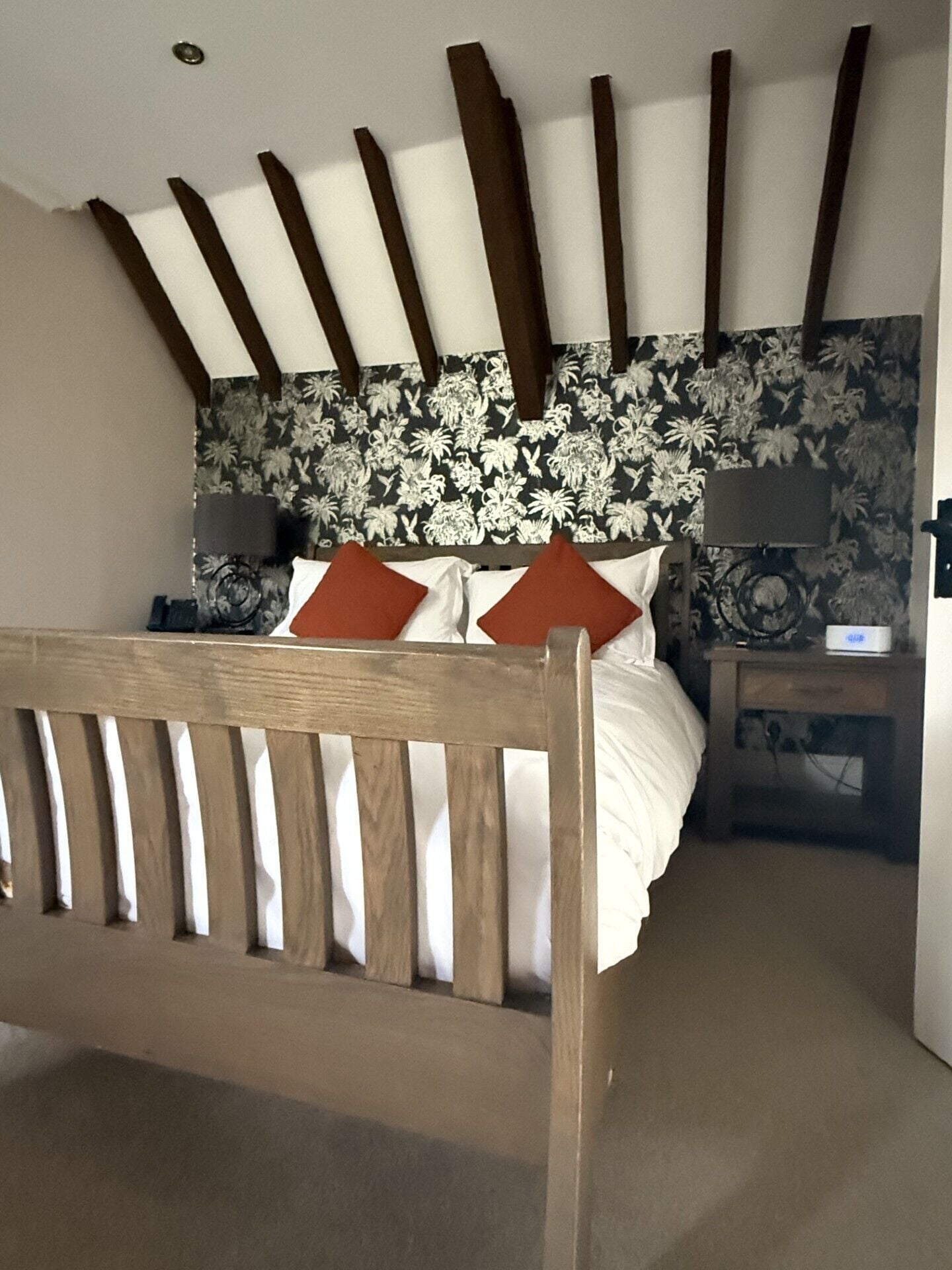 A bedroom with a wooden bed frame, a floral wallpaper headboard, and a nightstand with a lamp.