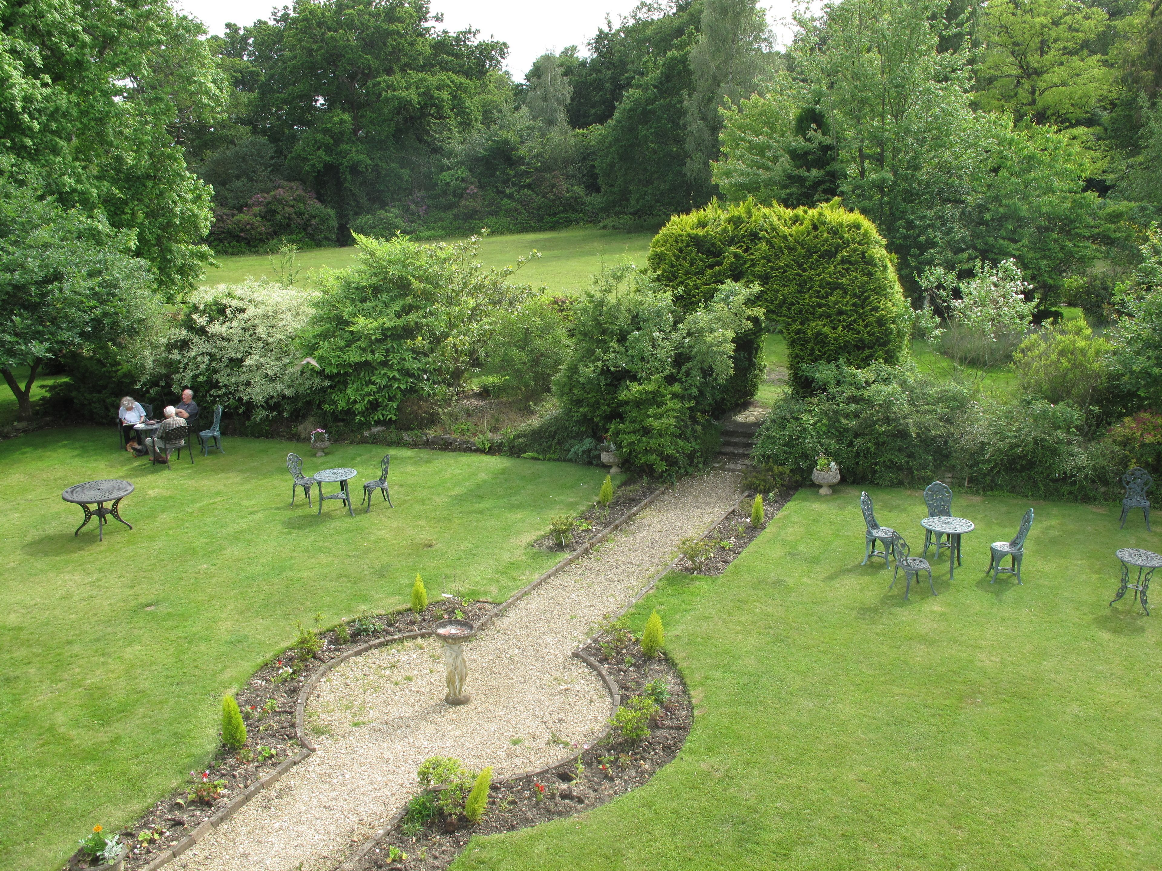 A garden with a central gravel path, surrounded by grass and various plants. There are several tables and chairs arranged for outdoor dining.
