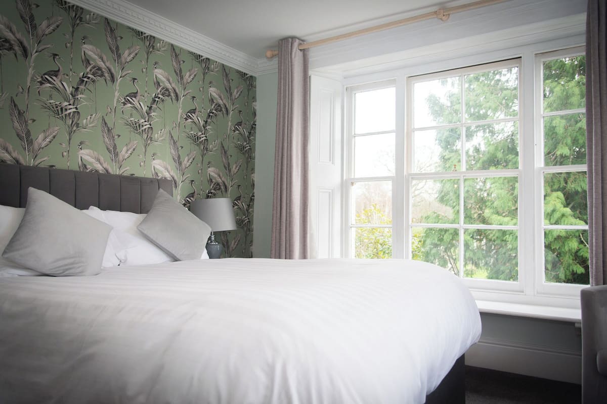 Una cama bien hecha con sábanas y almohadas blancas, una lámpara de noche y una ventana con vistas a árboles.