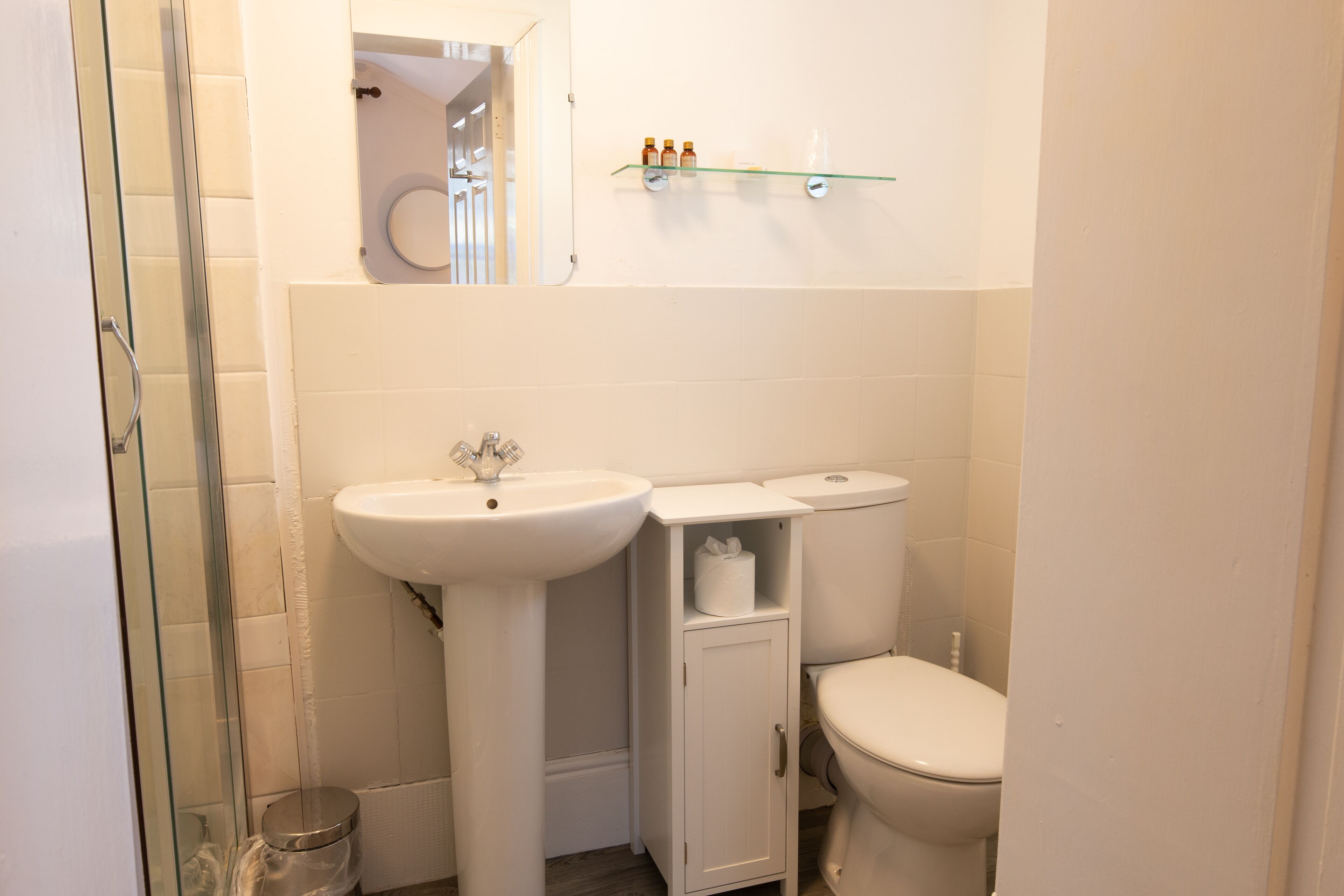 A small bathroom with a glass shower enclosure, a pedestal sink, a toilet, and a shelf with toiletries.
