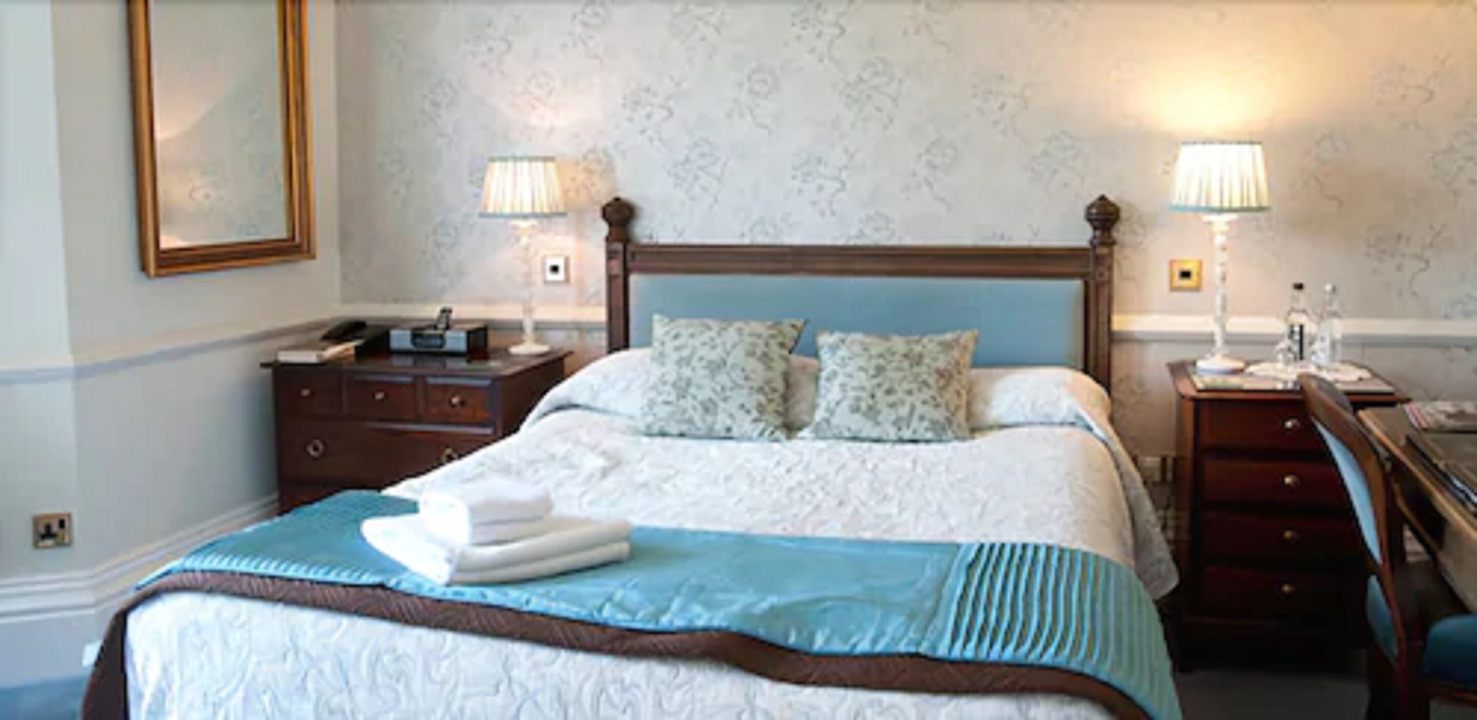 A neatly made bed with a blue and white bedspread, two pillows, and a folded towel on the side. There are two bedside tables with lamps and a small nightstand with a telephone.