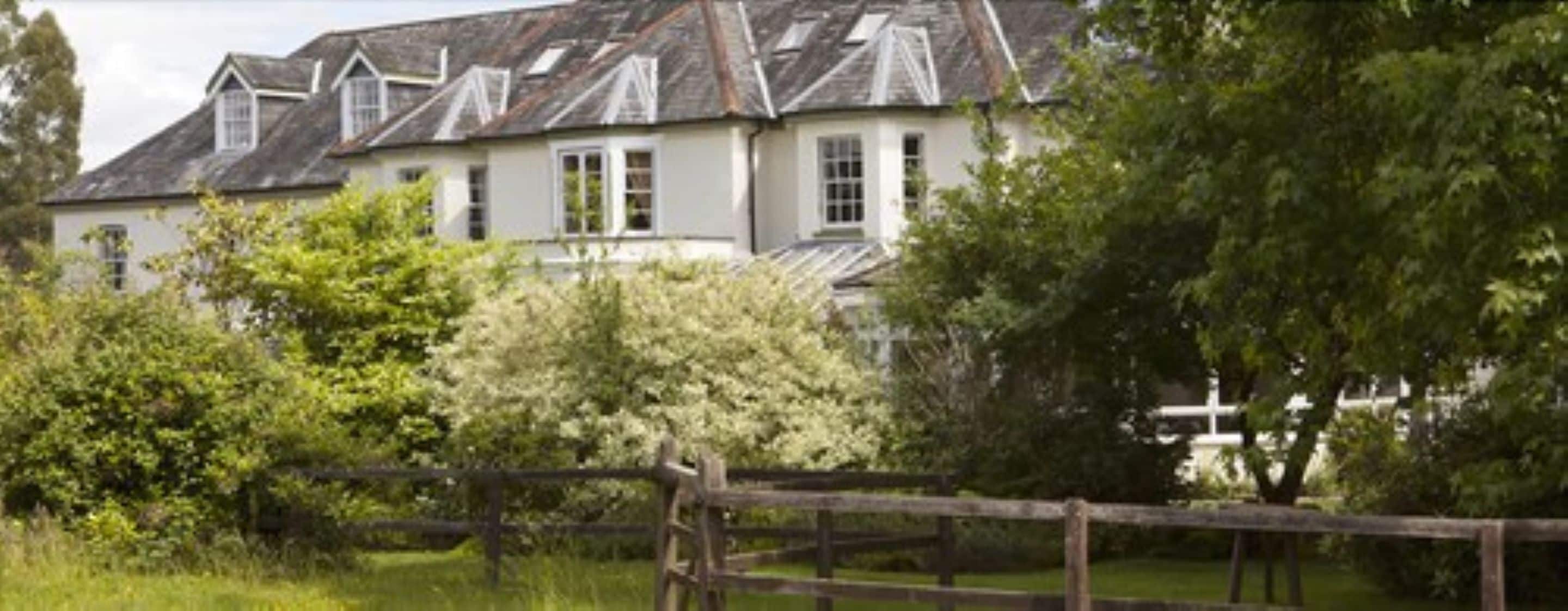 A large, white building with multiple windows and a sloped roof, surrounded by lush greenery and a wooden fence.