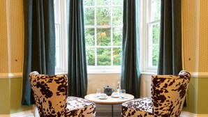 Una habitación con dos sillones estampados con vaca, una mesita redonda con tetera, tazas y vasos, y una ventana con cortinas.