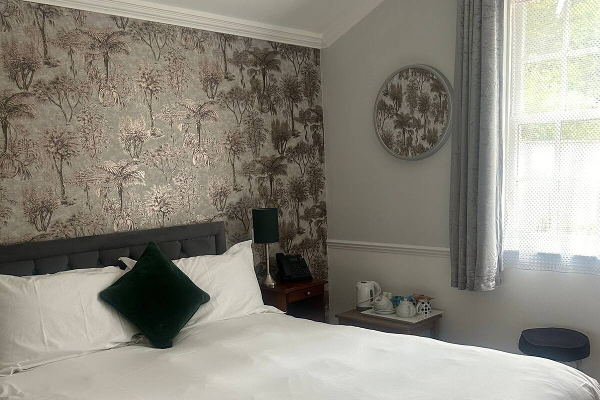 Una cama bien hecha con sábanas blancas y una almohada verde, una mesita de noche con una lámpara y un teléfono, una pared con estampado de papel tapiz floral, y una ventana con vistas a la vegetación.
