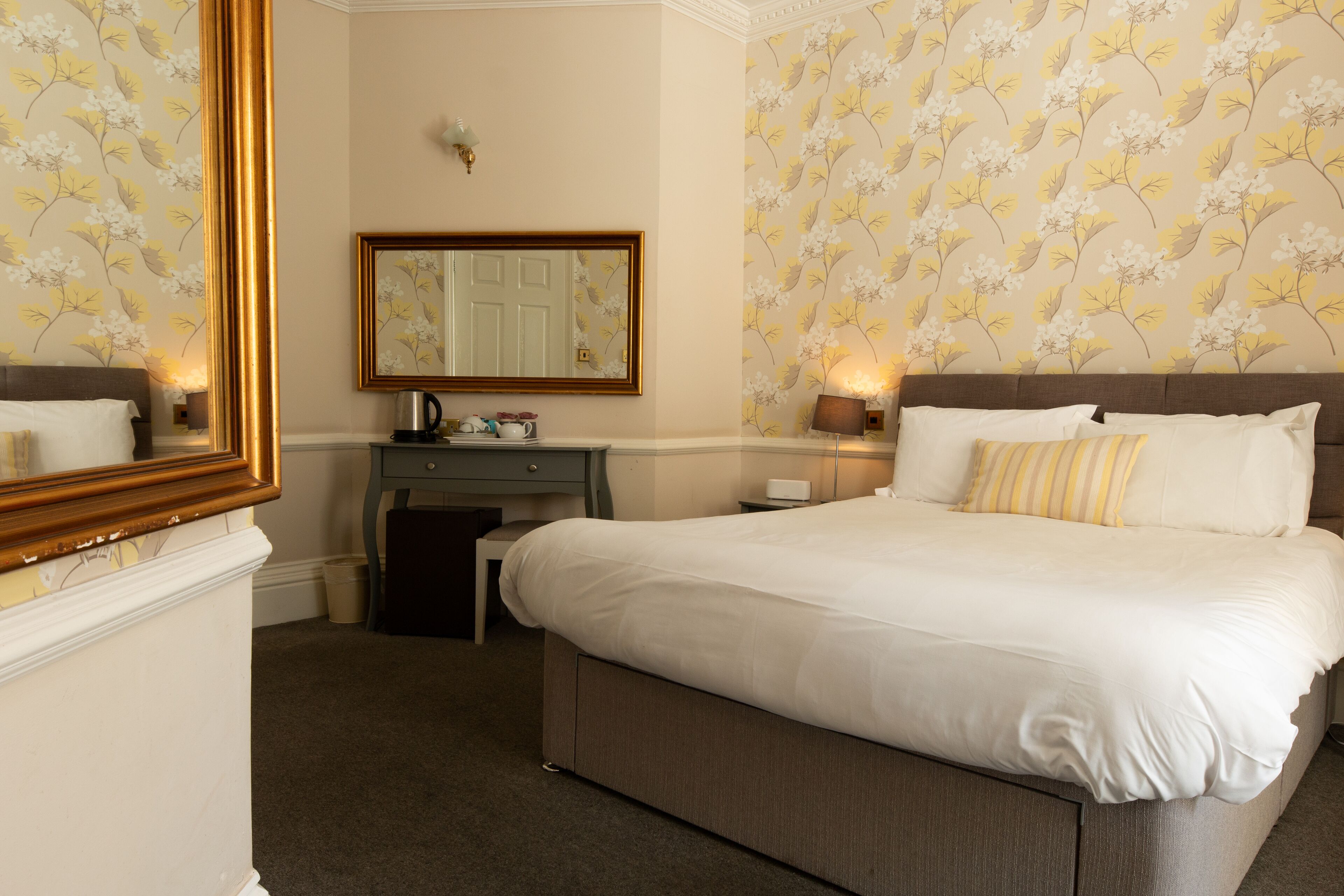 A hotel room with a large bed, a dresser, a mirror, and floral wallpaper.