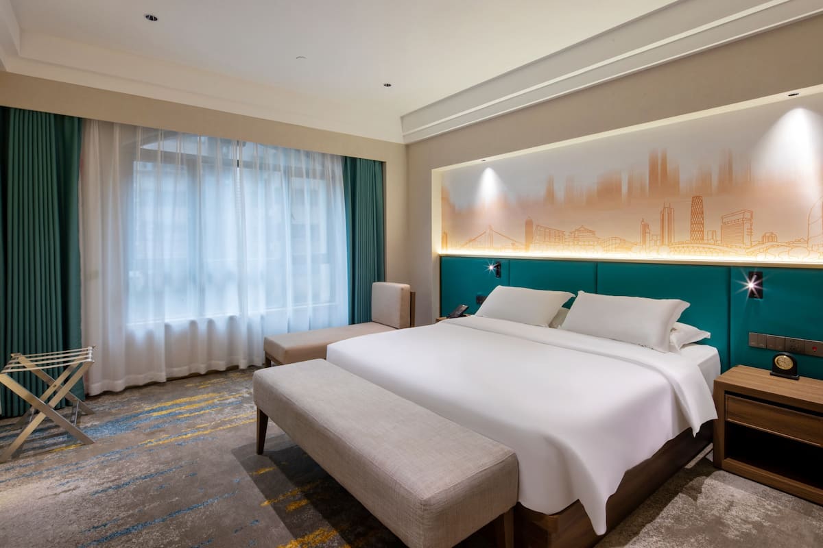 Een hotelkamer met een groot bed, een bankje, een nachtkastje en een stadsbeeld op de muur.