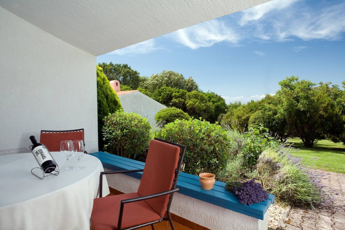 Een tafel met een wijnfles en glazen, een stoel en uitzicht op een tuin met bomen en een kiezelpad.