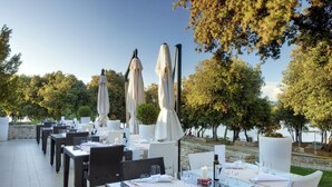 An outdoor dining area with tables and chairs, umbrellas, and a view of trees and water. - Valamar Tamaris Villas (Tar-Vabriga)