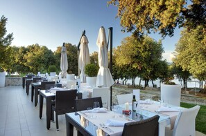 Een terras met tafels en stoelen, parasols, en uitzicht op bomen en water.