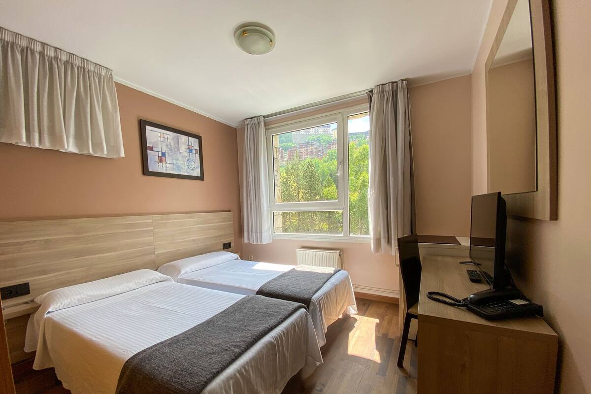 Habitación de hotel con dos camas, un escritorio con computadora y teléfono, una ventana con cortinas y un cuadro en la pared.
