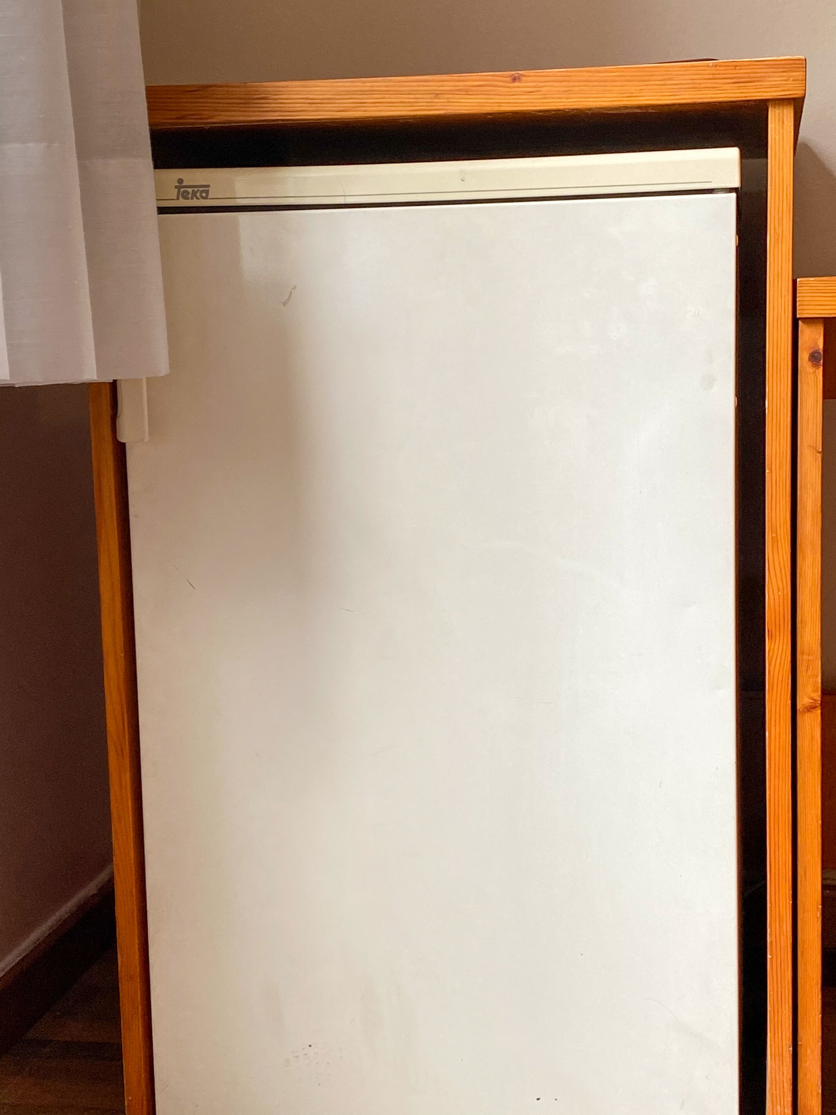 Un refrigerador blanco con estructura de madera, situado en una habitación con pared lisa y marco de puerta de madera.