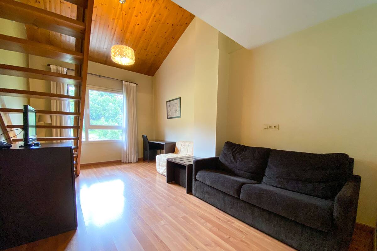Un espacio habitable amplio con techo de madera, un sofá, una mesita y un televisor sobre un soporte.