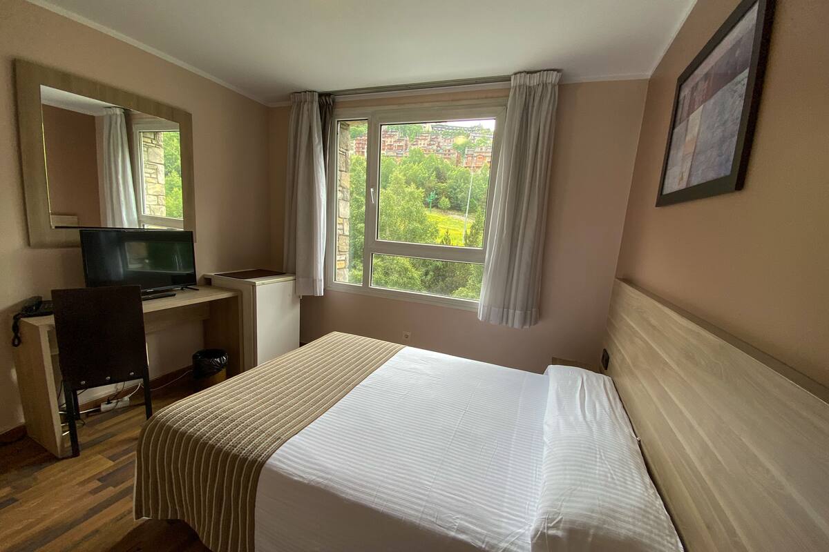 Una habitación de hotel con cama, escritorio con televisor, silla y ventana con cortinas.