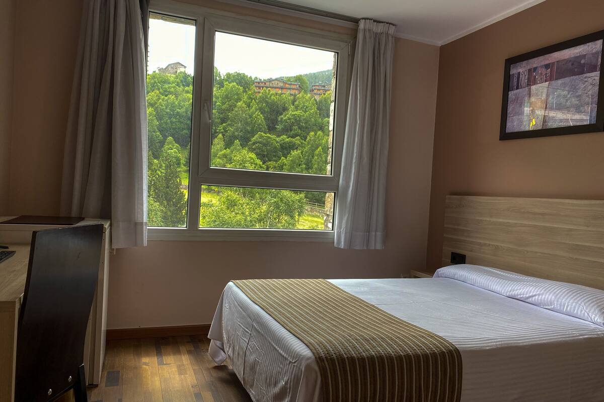 Habitación de hotel con cama, escritorio y ventana que ofrece vistas a zonas verdes y edificios.