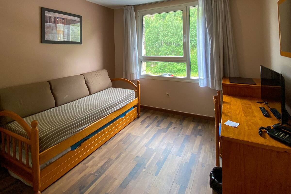 Habitación compacta con una cama de madera, un escritorio con computadora, una ventana con cortinas y un cuadro en la pared.