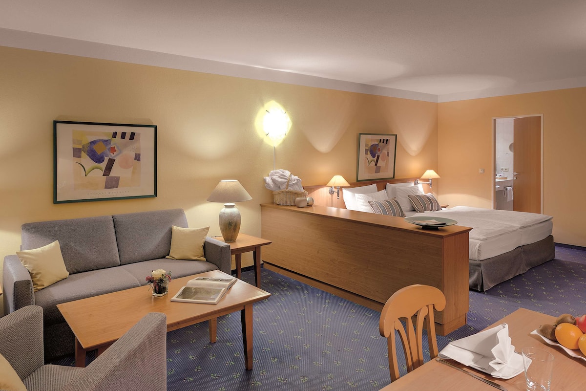 A hotel room with a sofa, a small table, a desk, a bed, and a breakfast table with fruit.