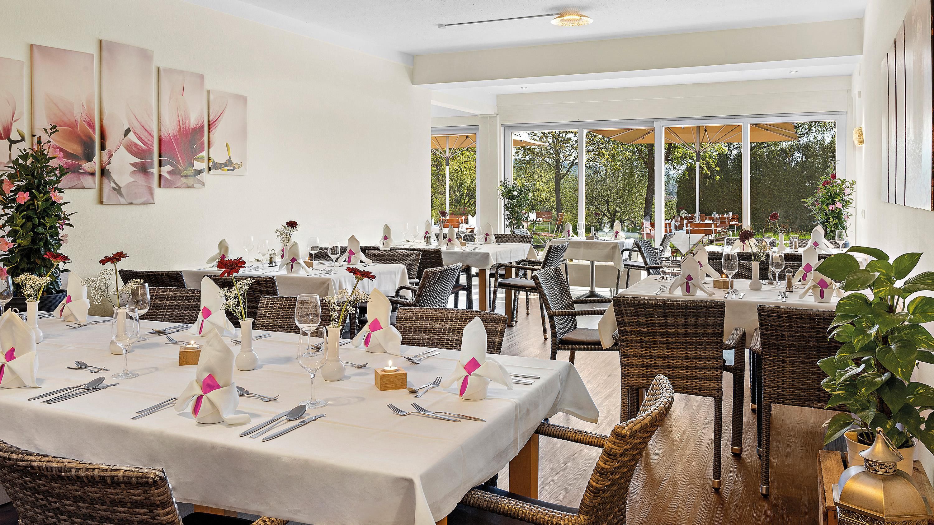 A restaurant with multiple tables set for dining, wicker chairs, and large windows offering a view of the outdoors.