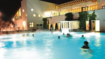 Ein Schwimmbad mit darin befindlichen Personen, umgeben von einem Gebäude mit zahlreichen Fenstern und einem überdachten Bereich mit Sonnenschirmen.