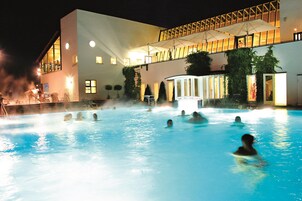 A swimming pool with people in it, surrounded by a building with multiple windows and a covered area with umbrellas.