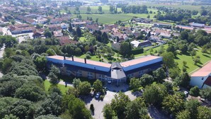 An aerial view of a residential area with a prominent building featuring a flat roof and large windows, surrounded by lush greenery and other houses. - Best Western Aparthotel Birnbachhoehe (Bad Birnbach)