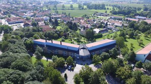 An aerial view of a residential area with a prominent building featuring a flat roof and large windows, surrounded by lush greenery and other houses. - Best Western Aparthotel Birnbachhoehe (Bad Birnbach)