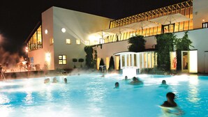 Zona de piscina con gente en el agua, un edificio con terraza cubierta y una entrada bien iluminada.
